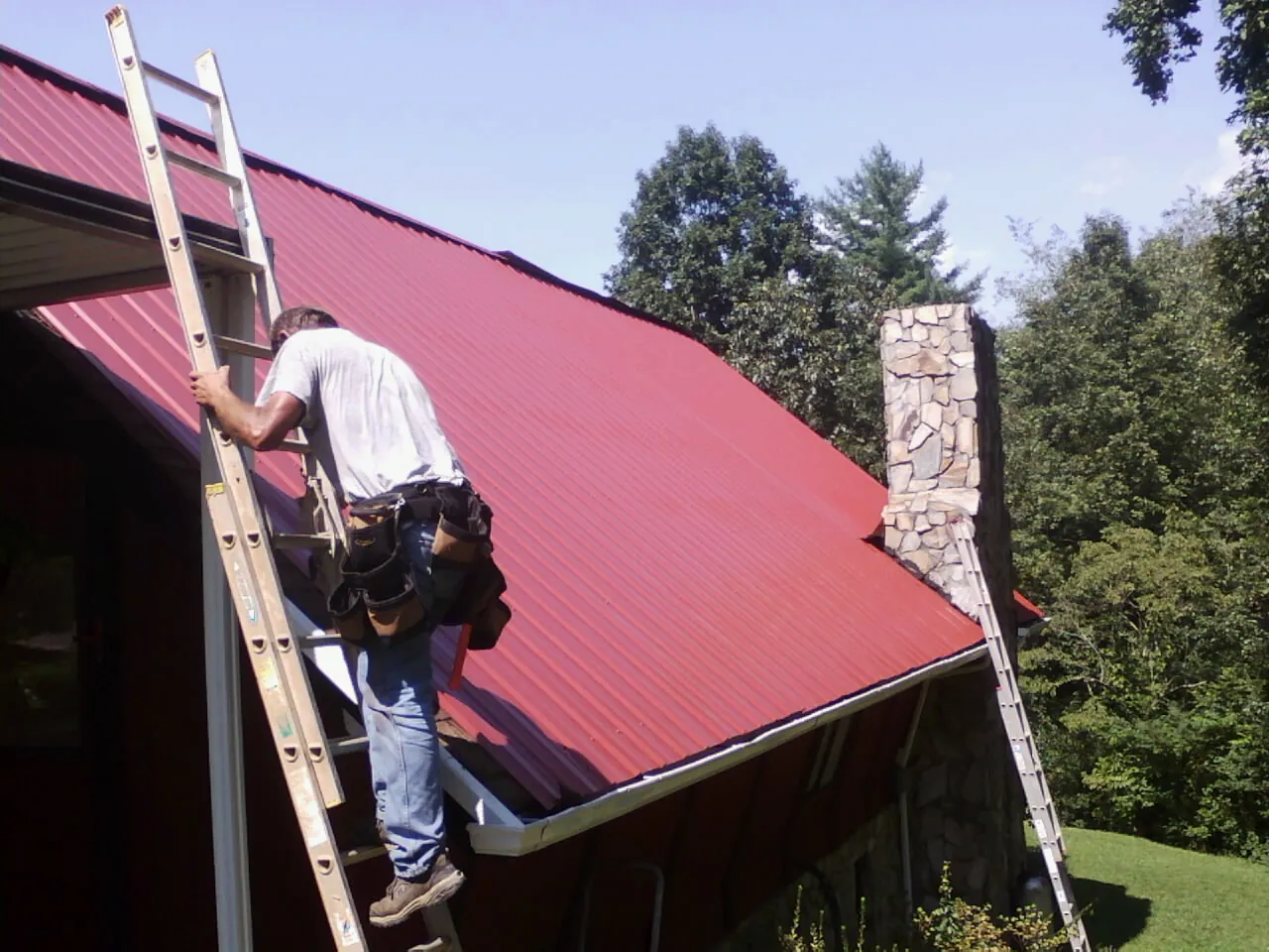 Roofer on a ladder beside a red metal roof and stone chimney — Soffit & Fascia Repair by Roofing Brothers in Shelly
